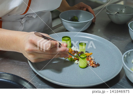 Chef meticulously plating modern gourmet cuisine, carefully arranging fresh ingredients with tweezers on a ceramic dish, creating a sustainable and healthy fine dining experience Chef meticulously plating modern gourmet cuisine, carefully arranging fresh ingredients with tweezers on a ceramic dish, creating a sustainable and healthy fine dining experience 141846975