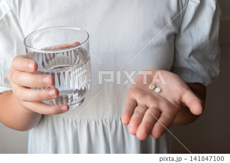 Girl in a dress take the two pills and glass with water in the hand. Healthcare from pharmacy. Kid sit at home because of illness. Girl in a dress take the two pills and glass with water in the hand. Healthcare from pharmacy. Kid sit at home because of illness. 141847100