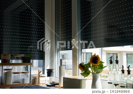 Exterior roller shutters partially cover large kitchen windows above a counter with jars, bottles and sunflowers Exterior roller shutters partially cover large kitchen windows above a counter with jars, bottles and sunflowers 141848969