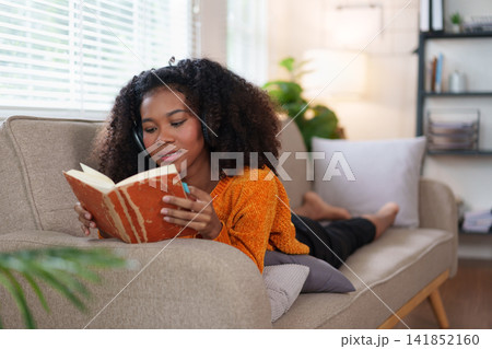 African American Woman Relaxing on Couch While Reading Book at Home. 141852160