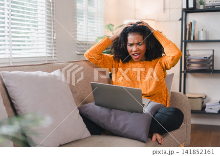 African American woman wearing headphones looks shocked while watching on laptop, sitting on couch at home. African American woman wearing headphones looks shocked while watching on laptop, sitting on couch at home. 141852161