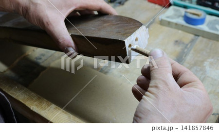 A close-up of a carpenter's hands inserting a wooden dowel into the end of a carved piece coated with PVA glue. The craftsman holds the piece over a workbench, preparing it for joining A close-up of a carpenter's hands inserting a wooden dowel into the end of a carved piece coated with PVA glue. The craftsman holds the piece over a workbench, preparing it for joining 141857846