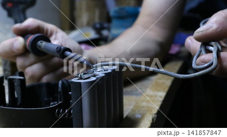 a lithium-ion battery being soldered. The technician holds a soldering iron in his left hand, pointing its tip toward the battery terminal. With his right hand, he applies thick tin solder to the join a lithium-ion battery being soldered. The technician holds a soldering iron in his left hand, pointing its tip toward the battery terminal. With his right hand, he applies thick tin solder to the join 141857847