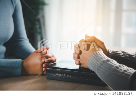 Two people praying over Holy Bible on wooden table with warm sunlight, representing Christian faith, hope, worship, spiritual support and peaceful devotion. Two people praying over Holy Bible on wooden table with warm sunlight, representing Christian faith, hope, worship, spiritual support and peaceful devotion. 141858157