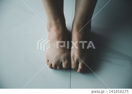 Close-up of bare feet placed together on a tiled floor, expressing nervousness, anxiety, insecurity, waiting, loneliness, and emotional vulnerability at home. Close-up of bare feet placed together on a tiled floor, expressing nervousness, anxiety, insecurity, waiting, loneliness, and emotional vulnerability at home. 141858275