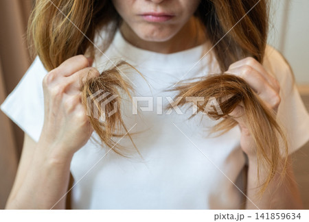Cropped shot view of Asian woman holding her damaged split ended hair. Hair damage is risk for further damage and breakage. It may also look dull or frizzy and be difficult to manage. Cropped shot view of Asian woman holding her damaged split ended hair. Hair damage is risk for further damage and breakage. It may also look dull or frizzy and be difficult to manage. 141859634