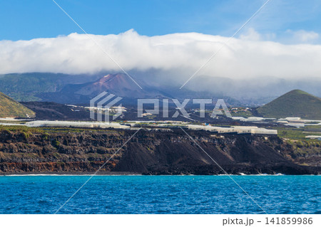 Volcanic cone of Tajogaite volcano visible on the horizon above clouds, shot from the sea near the lava delta in La Palma, Canary Islands, Spain Volcanic cone of Tajogaite volcano visible on the horizon above clouds, shot from the sea near the lava delta in La Palma, Canary Islands, Spain 141859986