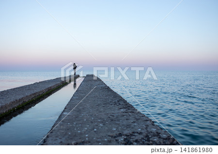 Long concrete pier extending into calm blue sea with a distant person at the end, emphasizing strong perspective, tranquil coastal scenery, horizon and minimalist seascape views Long concrete pier extending into calm blue sea with a distant person at the end, emphasizing strong perspective, tranquil coastal scenery, horizon and minimalist seascape views 141861980