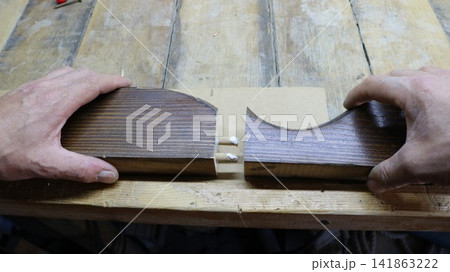A craftsman's hands join two shaped wooden pieces together on a workbench. Two dowels, coated with white PVA glue, protrude from the end of the left piece. The carpenter joins the pieces together. 141863222