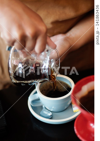 Fresh brewed black coffee being poured from a glass server into a white cup, capturing an authentic coffee preparation and serving scene with a warm cafe atmosphere. Fresh brewed black coffee being poured from a glass server into a white cup, capturing an authentic coffee preparation and serving scene with a warm cafe atmosphere. 141863436