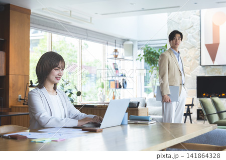 フリーアドレスのオフィスでデスク作業をする女性と奥に立つ男性社員 フリーアドレスのオフィスでデスク作業をする女性と奥に立つ男性社員 141864328