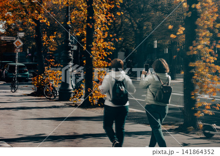 Two women walk through a tree-lined city street in autumn, one taking a photo with her phone as golden leaves glow in sunlight, capturing an everyday urban travel moment outside in warm autumn light. Two women walk through a tree-lined city street in autumn, one taking a photo with her phone as golden leaves glow in sunlight, capturing an everyday urban travel moment outside in warm autumn light. 141864352