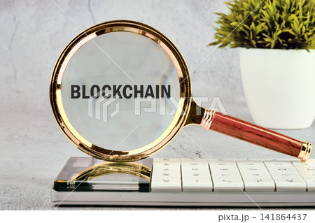 A magnifying glass highlights the word blockchain placed on a calculator. A small green plant adds to the scene A magnifying glass highlights the word blockchain placed on a calculator. A small green plant adds to the scene 141864437