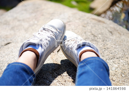 Close up woman leg and white sneakers sitting on the rock in natural sunlight Close up woman leg and white sneakers sitting on the rock in natural sunlight 141864598
