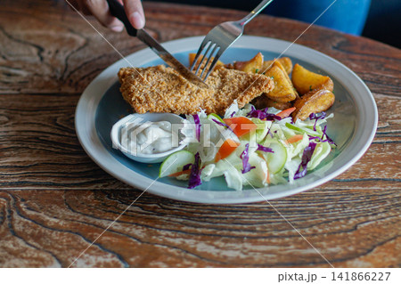 Close up of a person cutting a crispy breaded fish fillet served with fresh salad, potato wedges, and sauce on a rustic table. Close up of a person cutting a crispy breaded fish fillet served with fresh salad, potato wedges, and sauce on a rustic table. 141866227
