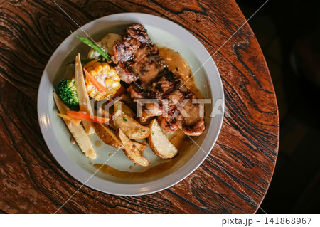 Grilled chicken with potato wedges, corn, broccoli, baby corn, carrots and brown gravy served on a white plate over a rustic wooden table, photographed from above as a restaurant meal. Grilled chicken with potato wedges, corn, broccoli, baby corn, carrots and brown gravy served on a white plate over a rustic wooden table, photographed from above as a restaurant meal. 141868967