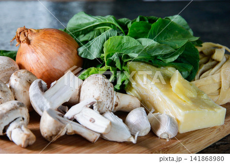 Fresh homemade pasta with mushrooms, leafy greens, onion, garlic and hard cheese arranged on a wooden board, illustrating Italian cooking, pasta preparation and fresh food ingredients. Fresh homemade pasta with mushrooms, leafy greens, onion, garlic and hard cheese arranged on a wooden board, illustrating Italian cooking, pasta preparation and fresh food ingredients. 141868980