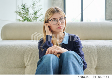 Sad teenage student girl in glasses sitting on floor hugging knees Sad teenage student girl in glasses sitting on floor hugging knees 141869834