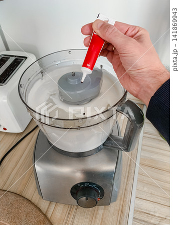 Hand adding red food coloring into a food processor with cream mixture preparation for cooking or baking in a kitchen stainless steel appliance on a wooden surface 141869943