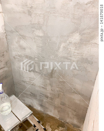 A grey cement wall with textured plaster finish stands next to a small white stepladder and a water spray bottle illustrating home renovation progress and interior decoration preparation in a room 141870018