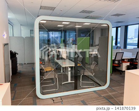 A modern glass meeting pod with chairs inside an open plan office creating a quiet space for collaboration in a corporate environment 141870139