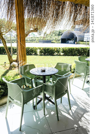 Outdoor cafe seating with green chairs and round tables on a sunny terrace, peaceful summer dining area surrounded by grass, trees and hedges Outdoor cafe seating with green chairs and round tables on a sunny terrace, peaceful summer dining area surrounded by grass, trees and hedges 141871360