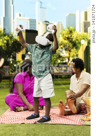Child playing with VR headset and raising both motion controllers while standing on picnic blanket, with parents relaxing nearby in sunny city park. Young boy using virtual reality gadget. 141871724