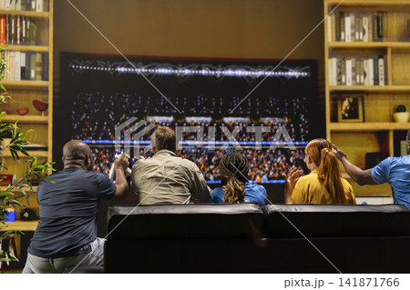Football supporters watch game on large television from couch in warm living room. Back view of soccer fans with tense body language following match on 4K TV screen Football supporters watch game on large television from couch in warm living room. Back view of soccer fans with tense body language following match on 4K TV screen 141871766