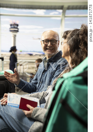 Confident older couple sitting together in airport terminal lounge, discussing travel plans and looking at departure screens. Breaking stereotypes about aging, traveling in later life. 141871778