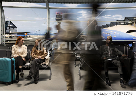 Smiling women talking in busy airport surrounded by multiethnic rushing blurry travelers. Sisters sitting on chairs waiting for plane and silhouette of people hurrying to fly home during holidays 141871779
