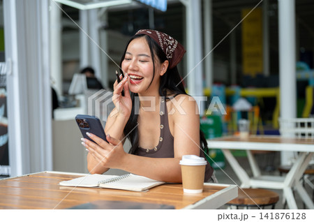 Young Asian woman using smartphone over notebook while relaxing with coffee at an outdoor cafe table 141876128