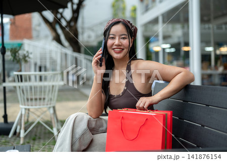 Young asian woman talking on phone and relaxing after shopping while sitting on bench at mall plaza. Young asian woman talking on phone and relaxing after shopping while sitting on bench at mall plaza. 141876154