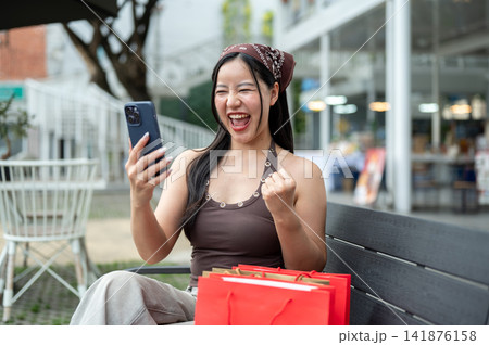 Excited asian woman shopper celebrating good news on phone with shopping bags on bench at mall plaza Excited asian woman shopper celebrating good news on phone with shopping bags on bench at mall plaza 141876158