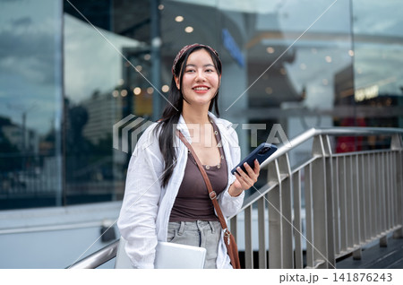 Young asian woman holding laptop with smartphone standing in front of a building in the modern city Young asian woman holding laptop with smartphone standing in front of a building in the modern city 141876243