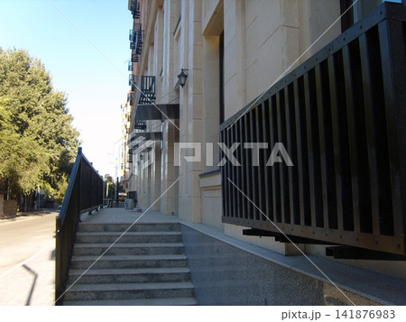 Modern multi-story building facade with light beige cladding, black metal balconies, and awnings. Steps lead from a street up to an entrance. Green trees are on the left under a blue sky. 141876983