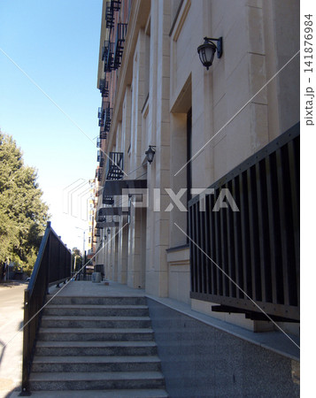 A view down a sidewalk with stone steps and black metal railings leading to a modern apartment building with beige stone facade, many balconies, awnings, and lanterns under a blue sky. 141876984