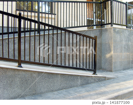 Outdoor ramp with grey stone slabs and black metal railings leading to a modern building entrance. Paved walkway in the foreground. 141877100