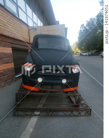 Custom matte black ZAZ-965 front body, featuring orange headlight trims and LED fog lights, displayed on a decorative metal stand in an urban street next to modern parked cars and buildings. 141879052