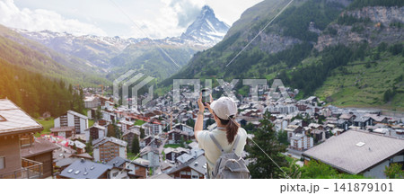 Woman Tourist Exploring Zermatt, Switzerland. Happy Traveler Taking Photo with Smartphone at Zermatt Village Viewpoint and Matterhorn. Popular Tourist Attraction and Travel Destination in Europe 141879101