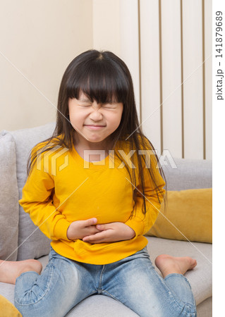 Asian little girl suffering stomach ache, sitting on sofa, painful expression, child health problem, abdominal pain concept Asian little girl suffering stomach ache, sitting on sofa, painful expression, child health problem, abdominal pain concept 141879689