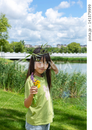 A young girl wearing a handmade willow garland and holding wildflowers smiles near a scenic riverbank. A young girl wearing a handmade willow garland and holding wildflowers smiles near a scenic riverbank. 141879999