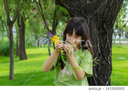 A young girl meticulously examines a bunch of colorful wildflowers while standing next to a large tree. 141880002
