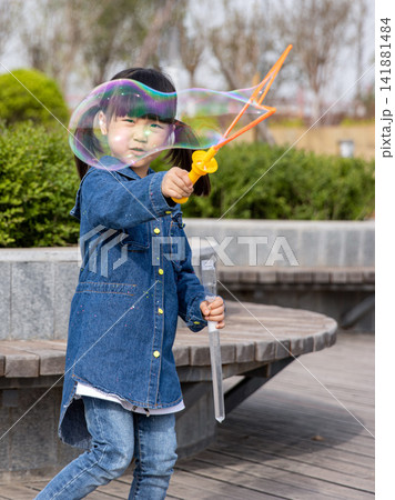 Joyful little girl holding a bubble wand and laughing with huge soap bubbles in the garden. 141881484
