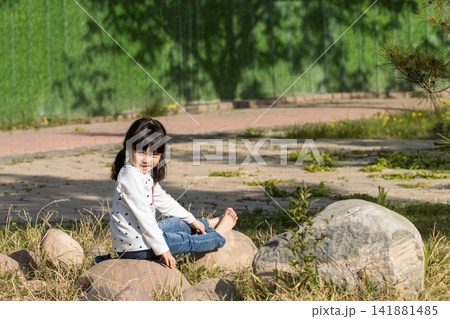 Small girl sitting alone on a large rock in the park, enjoying a quiet moment in nature. Small girl sitting alone on a large rock in the park, enjoying a quiet moment in nature. 141881485