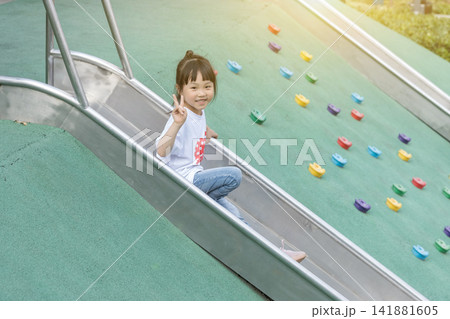 Joyful little girl sliding down a metal slide at a modern public playground. 141881605