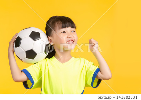 A high-energy portrait of the same East Asian girl on a solid yellow backdrop, holding the soccer ball aloft with one hand while cheering excitedly and raising her other fist in a victorious gesture, 141882331