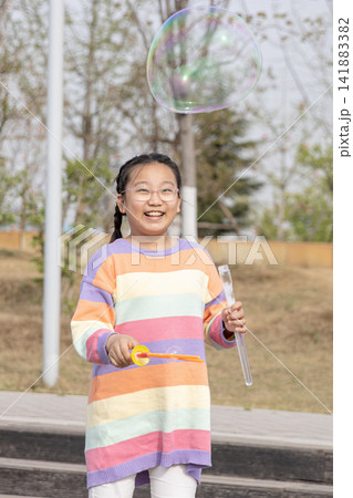 Portrait of a smiling young girl in a colorful sweater playing with a large soap bubble outdoors. Portrait of a smiling young girl in a colorful sweater playing with a large soap bubble outdoors. 141883382