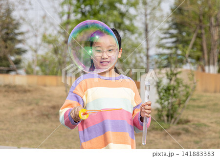 Portrait of a smiling young girl in a colorful sweater playing with a large soap bubble outdoors. Portrait of a smiling young girl in a colorful sweater playing with a large soap bubble outdoors. 141883383