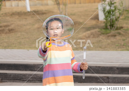 Portrait of a smiling young girl in a colorful sweater playing with a large soap bubble outdoors. Portrait of a smiling young girl in a colorful sweater playing with a large soap bubble outdoors. 141883385
