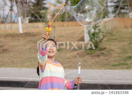 Portrait of a smiling young girl in a colorful sweater playing with a large soap bubble outdoors. 141883386